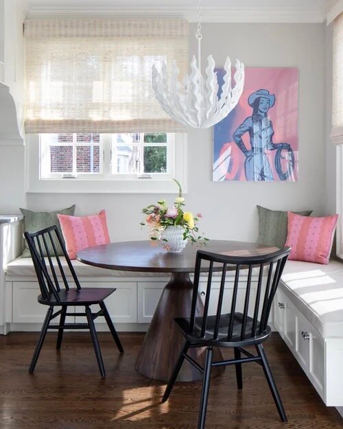 It Started with Wallpaper: Look Inside This Reimagined Richmond Colonial - 9 A round wooden table with two black chairs sits in a bright corner with built-in bench seating, pink and green pillows, a flower arrangement, and classic wallpaper that adds charm to this colorful Richmond home.