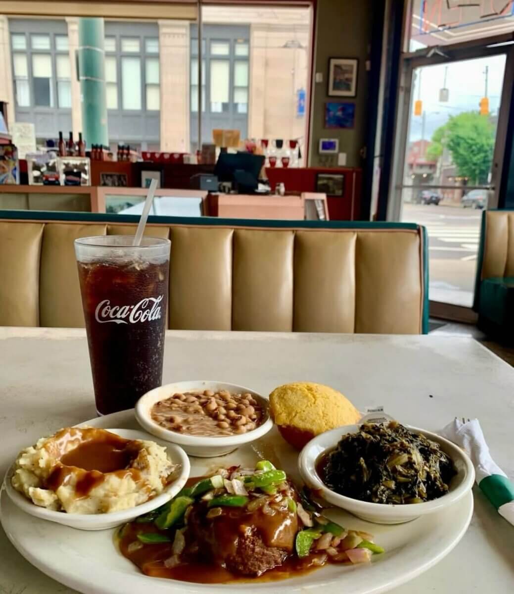 The South's Oldest Restaurants, Still Operating Today - 10 A plate with meatloaf, mashed potatoes with gravy, black-eyed peas, collard greens, cornbread, and a large Coca-Cola sits on a table in a diner with booths and large windows.