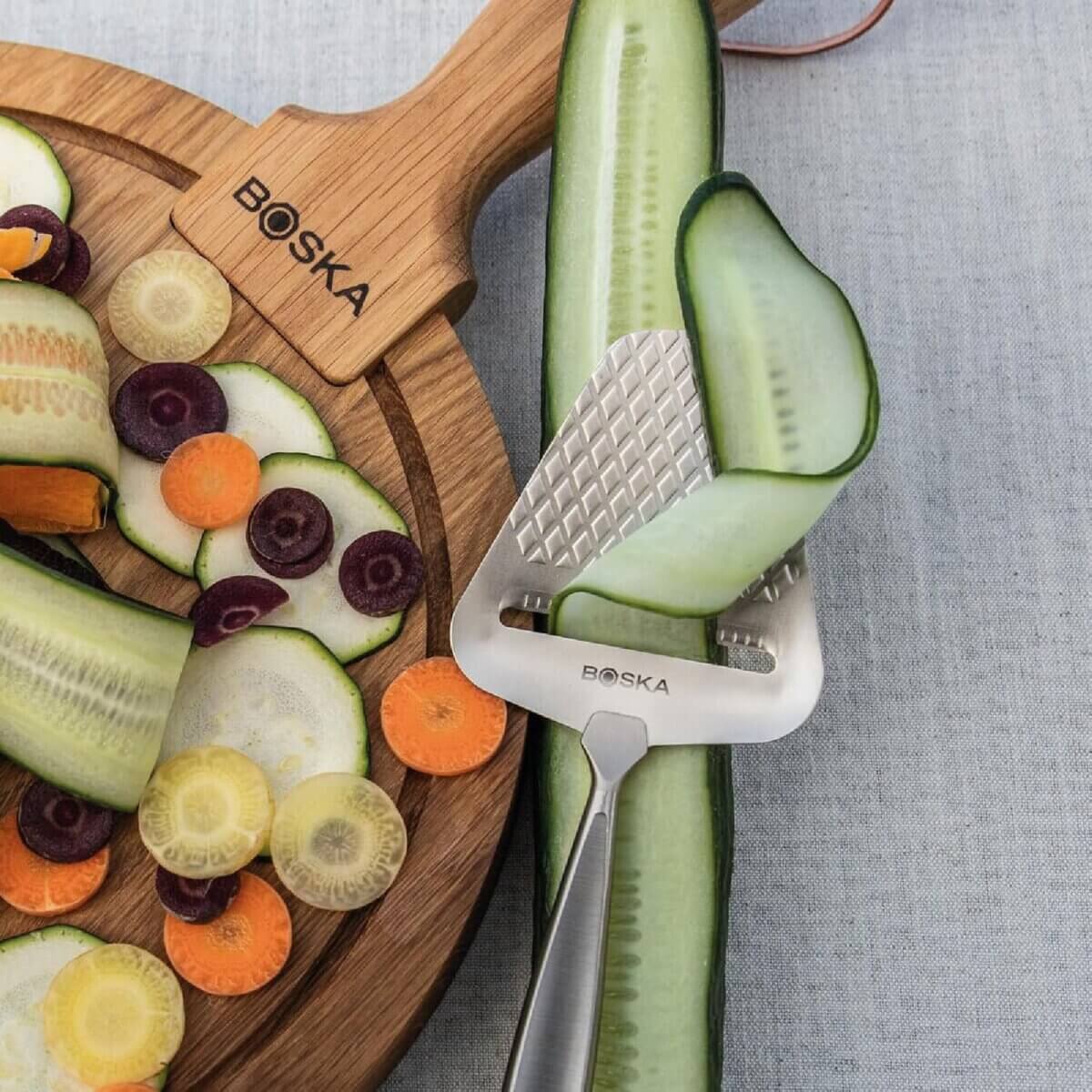 Chefs' Kitchen Tools Worth the Splurge (and Some to Save On) - 15 A metal vegetable peeler, a staple among chef tools, slices a thin strip from a cucumber, with assorted sliced vegetables on a wooden board in the background.
