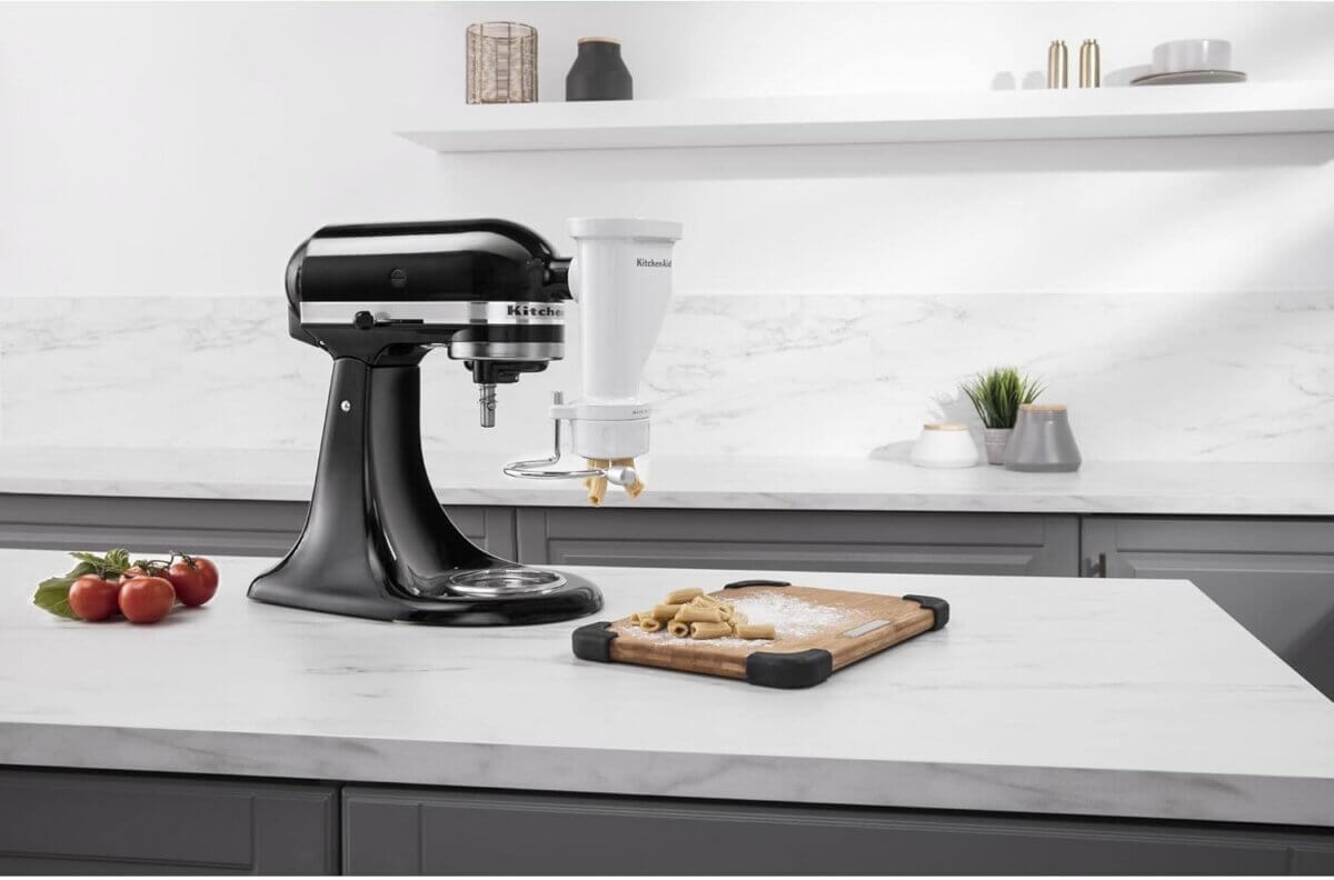 Chefs' Kitchen Tools Worth the Splurge (and Some to Save On) - 4 A black stand mixer, a staple among chef tools, sits on a kitchen counter with an attachment, next to a cutting board holding pasta dough and some tomatoes.