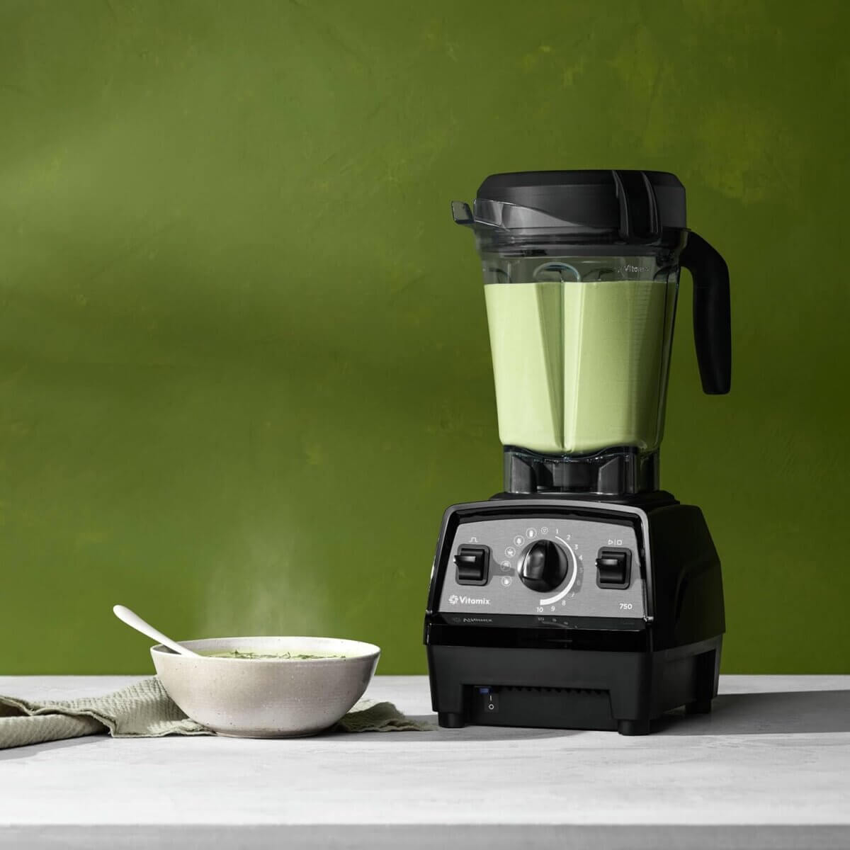 Chefs' Kitchen Tools Worth the Splurge (and Some to Save On) - 2 A black Vitamix blender, a staple among chef tools, is filled with green soup on a countertop next to a steaming bowl with a spoon, set against a green background.