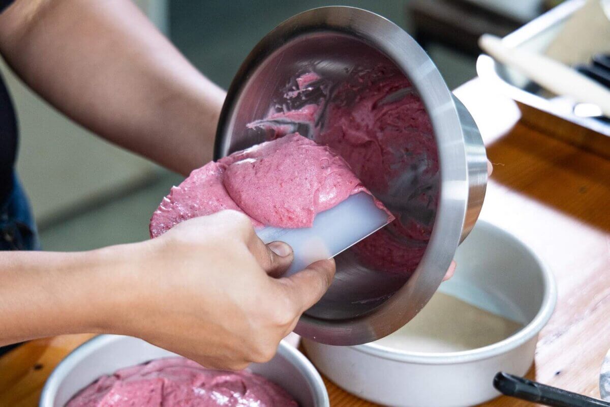 Chefs' Kitchen Tools Worth the Splurge (and Some to Save On) - 12 Using chef tools, a person uses a spatula to transfer pink cake batter from a metal bowl into a round cake pan on a wooden surface.