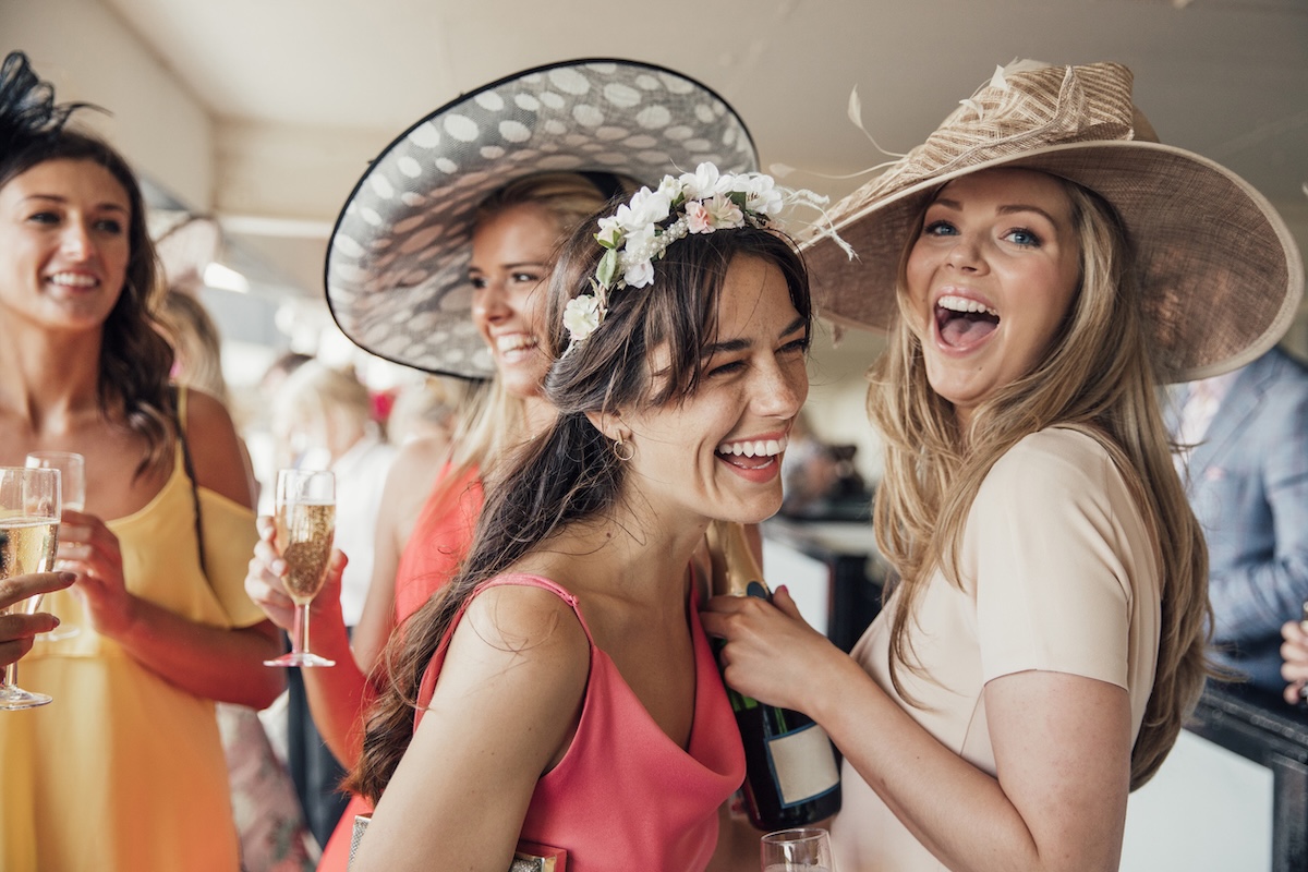 Four women dressed in colorful Kentucky Derby outfits and hats smile and laugh while holding drinks at a lively indoor social event.