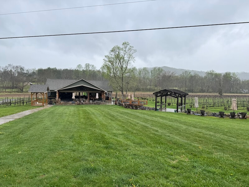 Spend a Weekend 'Wine-Hopping' in Asheville, NC - 12 Large grassy lawn leads to a covered outdoor pavilion, with vineyard rows and trees set against a cloudy sky—perfect for a relaxing weekend getaway near Asheville, NC.