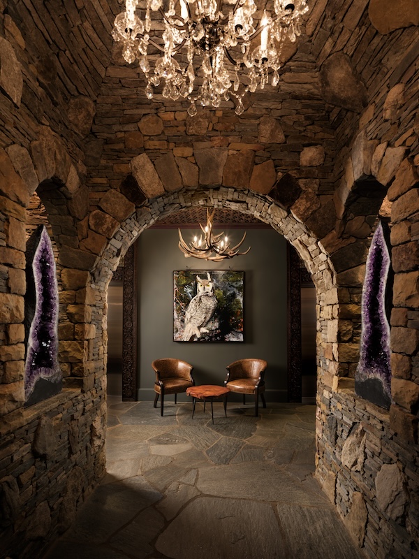 Spend a Weekend 'Wine-Hopping' in Asheville, NC - 3 Stone hallway with arched entry, crystal geodes on either side, chandelier above, and a seating area with two chairs, a round table, and an owl painting—perfect for relaxing after a weekend of winery hopping in Asheville NC.