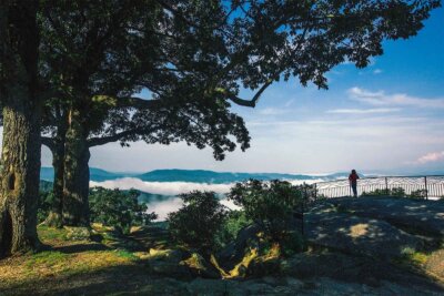 A person stands at a fenced viewpoint on a rocky hilltop near Asheville NC, overlooking distant mountains and clouds, with large trees framing the scene under a blue sky—perfect for a weekend getaway.
