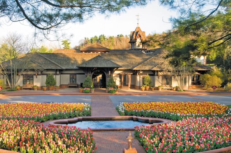 Spend a Weekend 'Wine-Hopping' in Asheville, NC - 7 A large building with a shingled roof and arched entrance stands behind a circular flower bed of colorful tulips and a central fountain—an inviting scene for a weekend getaway in Asheville NC. Trees and shrubs gracefully border the area.