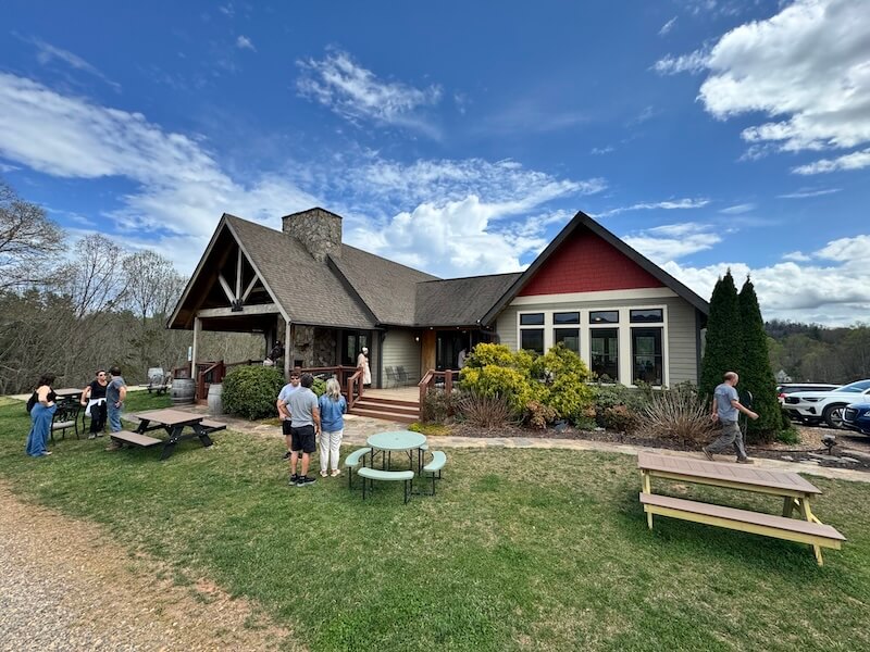Spend a Weekend 'Wine-Hopping' in Asheville, NC - 9 A group of people stand and talk outside a house with large windows, a stone chimney, and a porch, enjoying a weekend getaway near Asheville NC, surrounded by picnic tables and greenery under a partly cloudy sky.