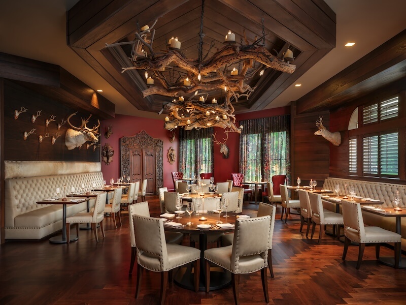 Spend a Weekend 'Wine-Hopping' in Asheville, NC - 4 Elegant restaurant dining room with beige upholstered chairs, round tables set for meals, mounted animal heads, wood accents, and a chandelier made of branches with candles—perfect for a weekend escape or winery hopping near Asheville NC.