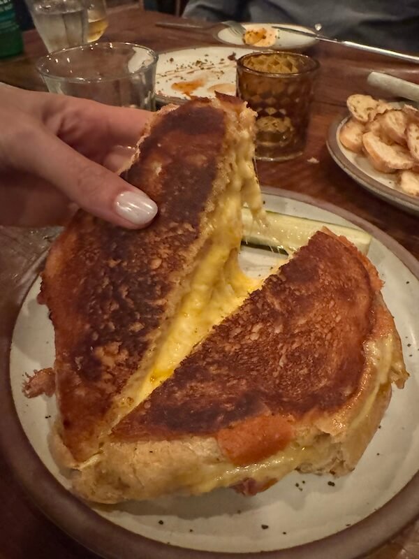 Spend a Weekend 'Wine-Hopping' in Asheville, NC - 11 A hand pulls apart a grilled cheese sandwich on a plate, showing melted cheese inside—a perfect snack between winery hopping adventures near Asheville NC. Glasses and a plate of sliced bread are in the background, enhancing this cozy weekend vibe.