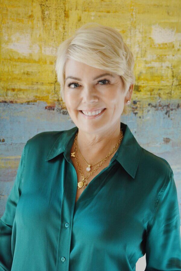 Behind the Nashville Symphony Fashion Show with Marci Houff - 2 Marci Houff wears a teal blouse and gold necklaces, and stands smiling in front of a yellow and blue abstract background.