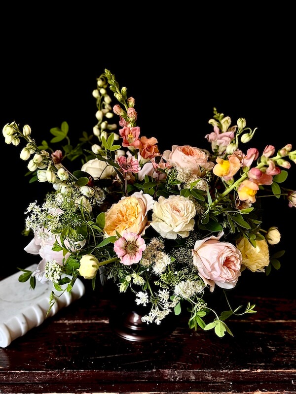 Meet the Nashville Grower Behind Bloom & Bounty - 5 A floral arrangement in a dark vase by Meredith Bishop features pale pink, peach, and yellow flowers with greenery, set against a dark background on a wooden surface.