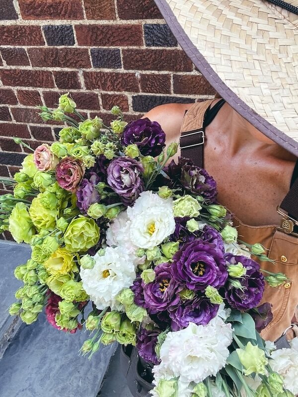 Meet the Nashville Grower Behind Bloom & Bounty - 4 Person wearing a wide-brimmed hat, face partly hidden, holds a large bouquet of purple, white, and green flowers while standing in front of a brick wall.