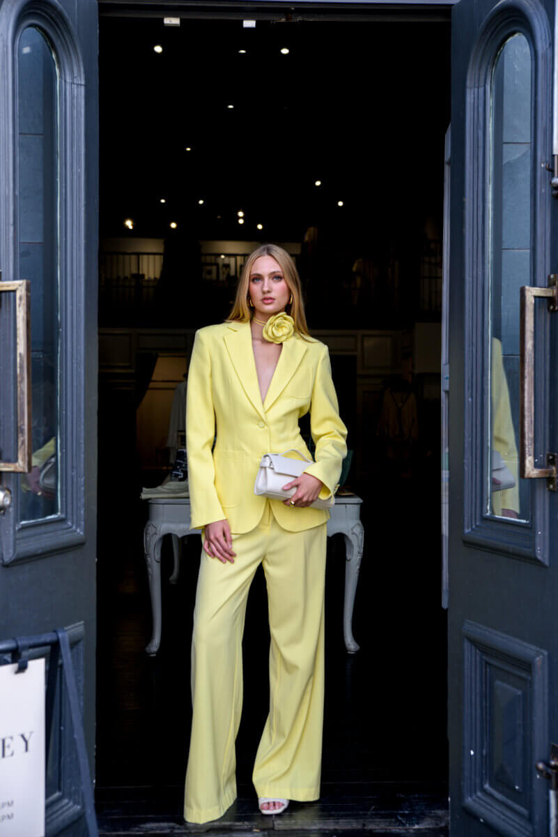 What to Wear to Spring Events: Party-Perfect Looks from Nashville Boutiques - 12 A woman stands in a doorway wearing a yellow suit with wide-leg pants and a floral accessory, holding a small white purseβan elegant look inspired by Nashville Boutiques.