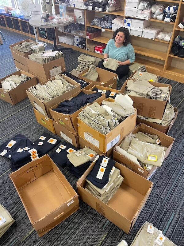 It Started with a StyleBlueprint Story — Now This Nashville Nonprofit Is Turning 10! - 3 A person sits on the floor surrounded by large boxes filled with neatly folded khaki pants and shorts in a room with shelves holding clothing and supplies, reflecting strong Nashville community connections.