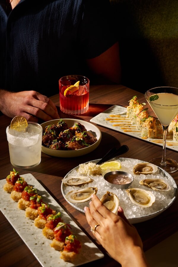 A table set with oysters on ice, sushi rolls, chicken wings, and cocktails at one of the new Nashville restaurants; two people are seated, one reaching for an oyster.