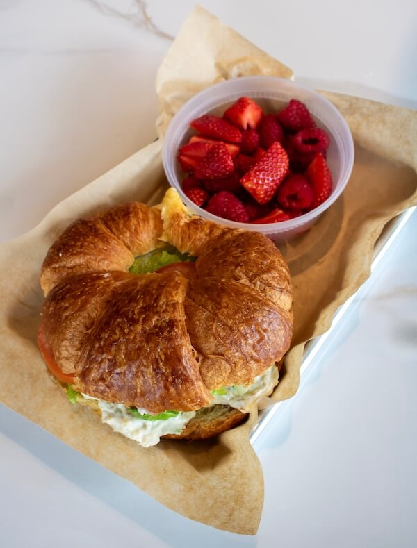 A croissant sandwich with lettuce and a plastic container of sliced strawberries and raspberries on brown parchment paper in a tray, inspired by flavors from new Nashville restaurants opening April 2026.