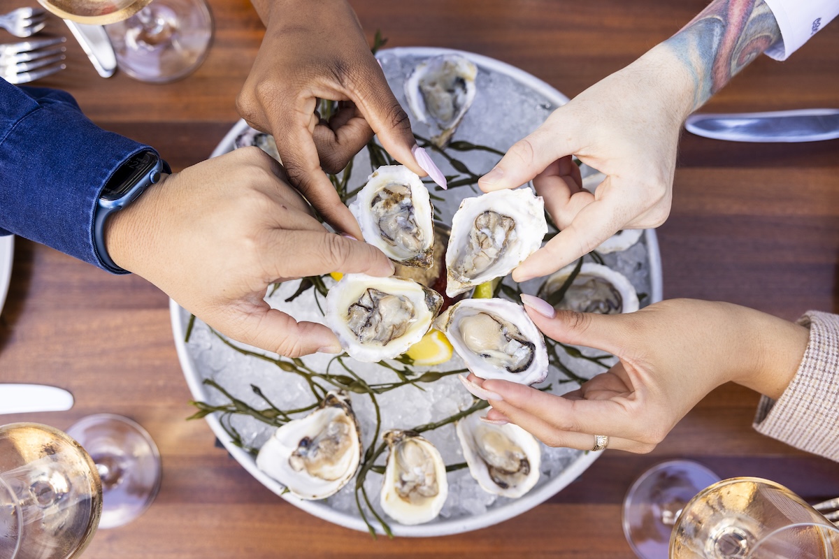Four hands holding fresh oysters over a plate of oysters on ice with lemon slices, on a wooden table set for dining—an inviting scene at one of the top new Nashville restaurants opening April 2026.