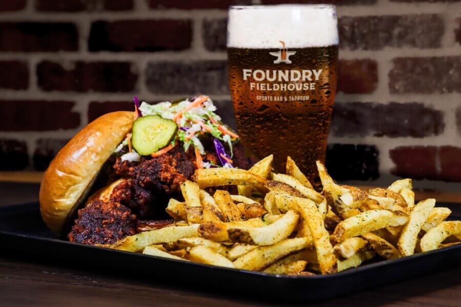 A plate with a fried chicken sandwich topped with slaw and pickles, seasoned fries, and a pint of beer in a Foundry Fieldhouse glass on a table—perfect for fans of new Nashville restaurants opening in April 2026.