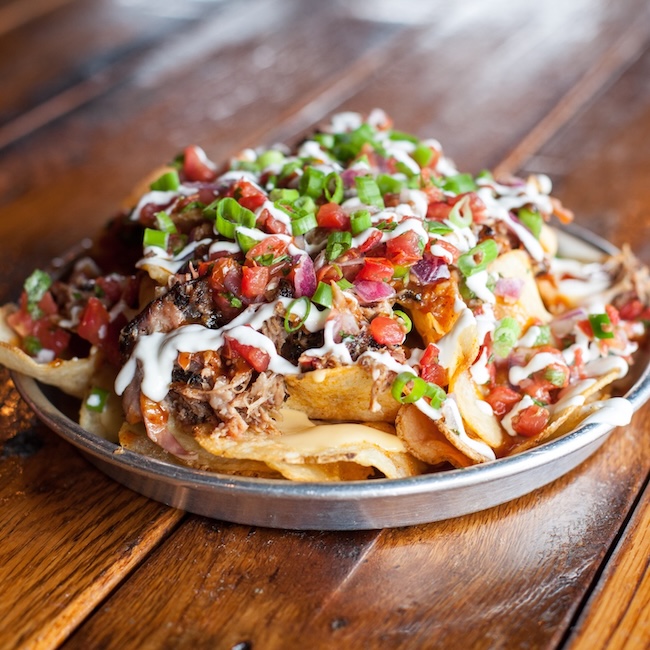 A metal plate of loaded nachos topped with shredded meat, diced tomatoes, green onions, cheese sauce, and drizzled with sour cream sits on a wooden table—a must-try at the new Nashville restaurants opening April 2026.