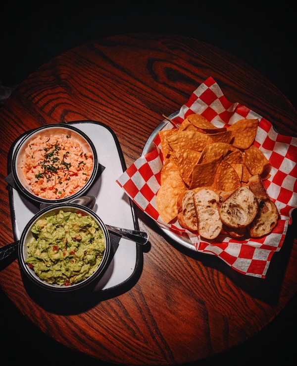 A wooden table with a tray of guacamole and cheese dip, served with tortilla chips and toasted bread on red and white checkered paper, perfect for sampling the flavors of Nashville restaurants in April 2026.