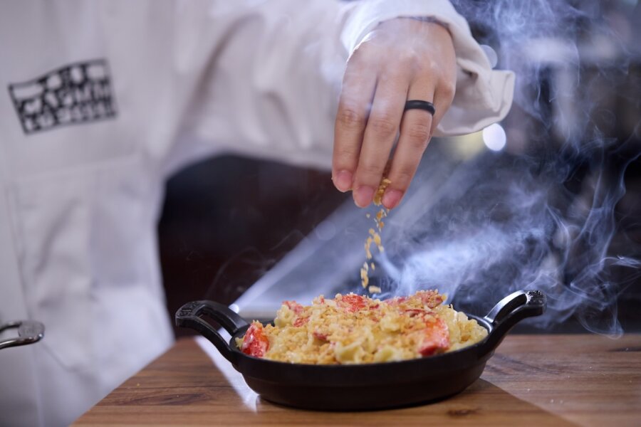 A person in a chef's jacket sprinkles seasoning onto a steaming lobster macaroni and cheese in a black skillet, showcasing the creative flair found in new Nashville restaurants debuting April 2026.