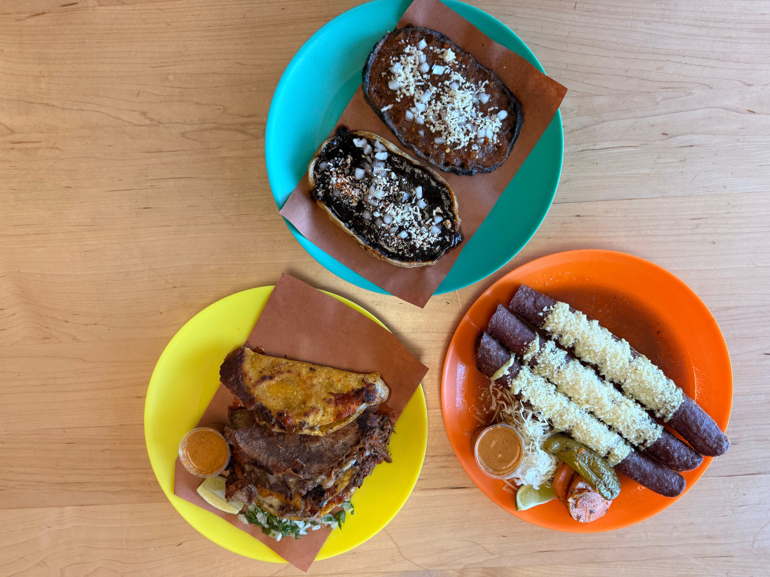 Three colorful plates with Mexican dishes at one of the new Nashville restaurants: two blue corn sopes with toppings, a stack of carnitas tacos, and four enchiladas topped with cheese and sauce. All on a wooden table.
