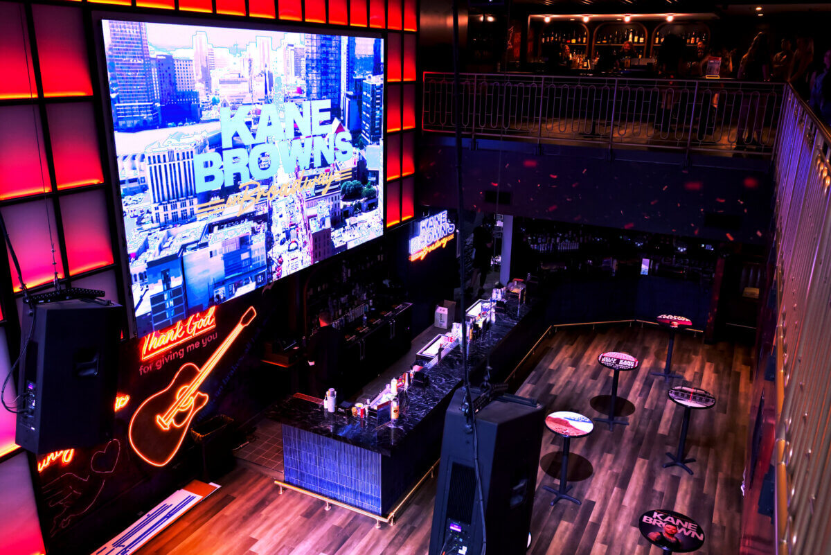 11 New Nashville Restaurants to Try This Month - 7 Bar interior with neon lights, a large screen displaying "Kane Brown," a guitar graphic, and a modern counter setup—plus an elevated balcony seating area above, capturing the vibrant energy of new Nashville restaurants.