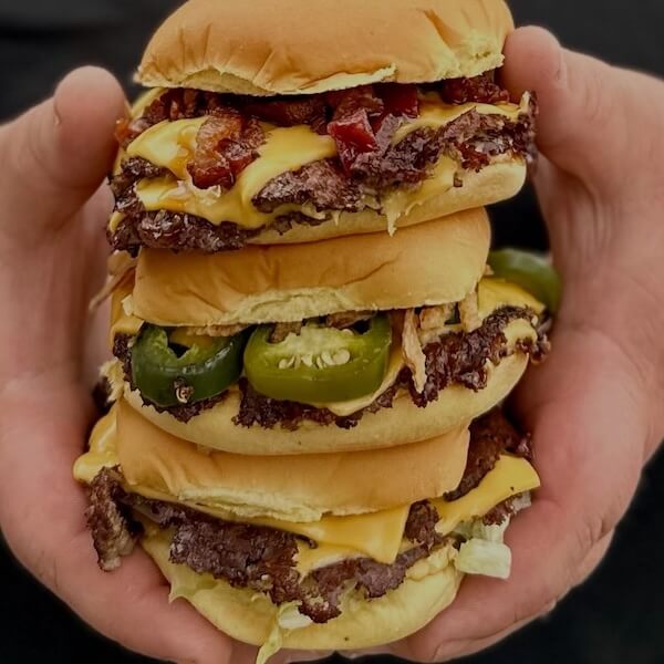 10 Nashville Smashburgers Worth the Hype - 2 A person holds three stacked smashburgers with toppings like bacon, jalapeños, and melted cheese in their hands.