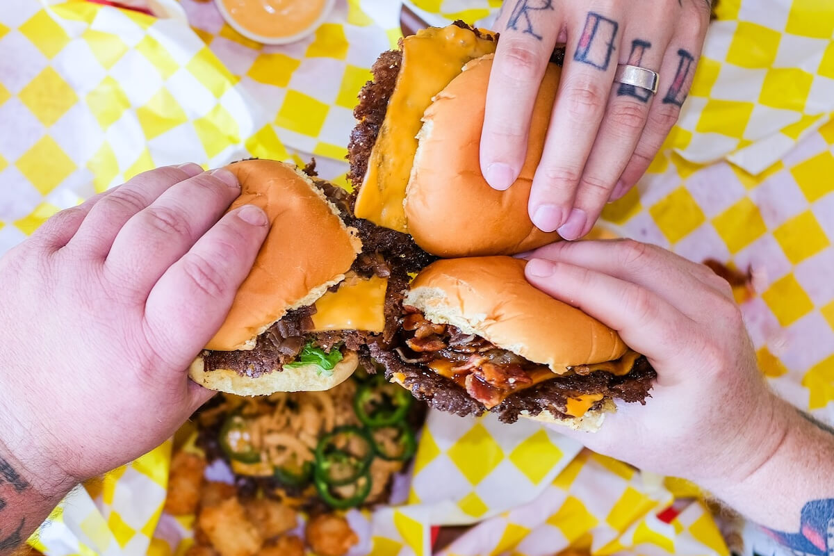 Three hands holding hype-worthy cheeseburgers over checkered yellow and white paper, with tater tots, jalapeños, and dipping sauce in the background—these Nashville smashburgers are among the best burgers Nashville has to offer.