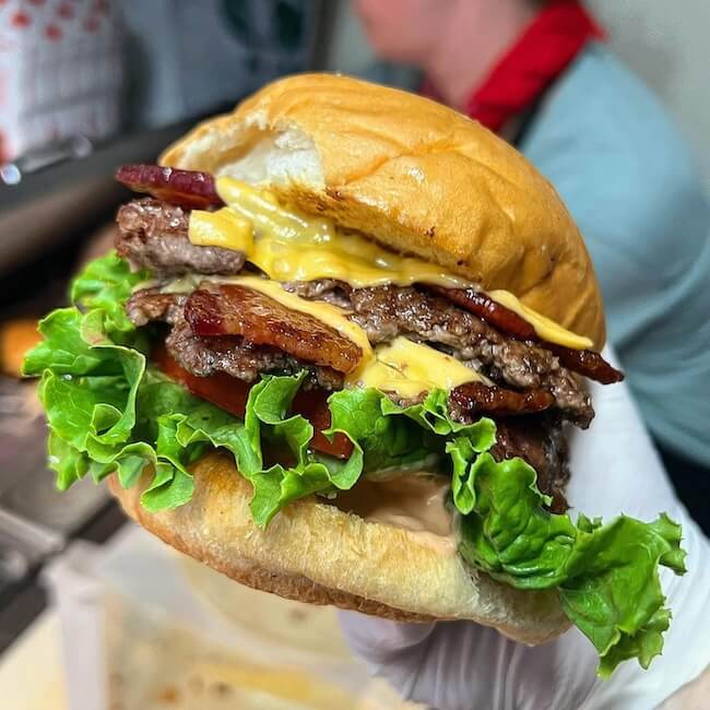 10 Nashville Smashburgers Worth the Hype - 5 A hand holds a Nashville Smashburger stacked with lettuce, tomato, bacon, two beef patties, melted cheese, and sauce in a soft bun.