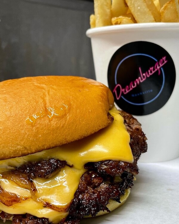 10 Nashville Smashburgers Worth the Hype - 4 A cheeseburger with melted cheese and grilled onions in a bun sits in front of a cup of French fries labeled "Dreamburger," serving up one of Nashville’s best burgers for true smashburgers fans.