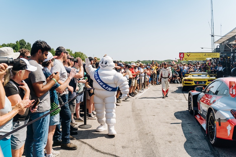 Meet Virginia Raceway Queen Connie Nyholm - 5 A crowd watches as the Michelin Man mascot walks in the pit lane beside parked sports cars at a racing event.