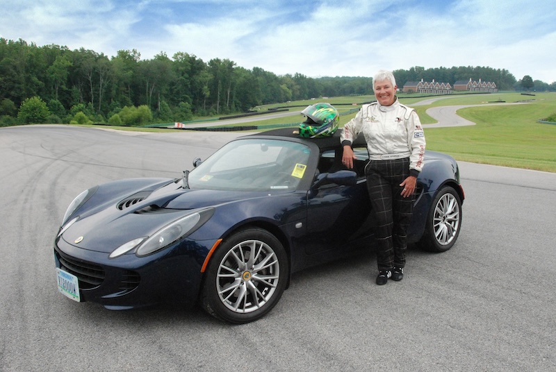 Meet Virginia Raceway Queen Connie Nyholm - 4 Connie Nyholm, in a racing suit, stands next to a dark convertible sports car on a racetrack, with trees and buildings in the background. A helmet rests on the car's roof.