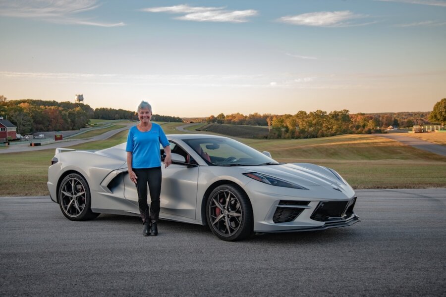 Meet Virginia Raceway Queen Connie Nyholm - 2 A woman, known as the Raceway Queen, stands next to a white sports car parked on a paved road with a racetrack and trees in the background.