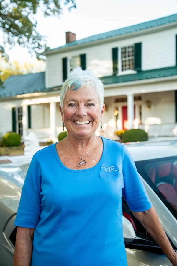 Meet Virginia Raceway Queen Connie Nyholm - 6 Connie Nyholm, an older woman with short gray hair, smiles warmly while wearing a blue shirt and standing in front of a white house with green shutters and a car.