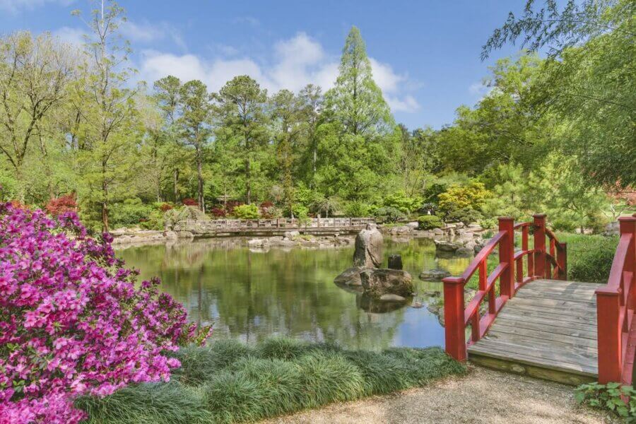 Your Guide to Mountain Brook's Villages - 10 A wooden red bridge leads to a pond surrounded by lush greenery, flowering bushes, and trees under a blue sky in Mountain Brook Villages.