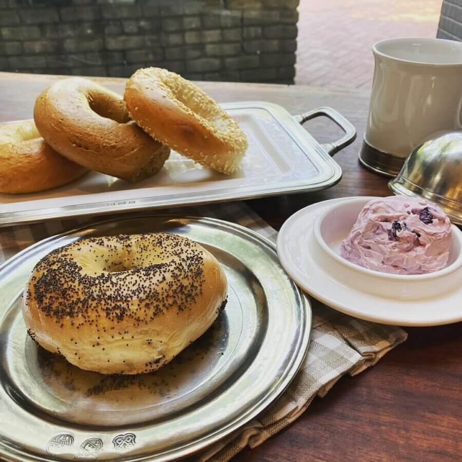 Your Guide to Mountain Brook's Villages - 11 A poppy seed bagel on a metal plate sits beside berry cream cheese, with plain and sesame bagels and a mug in the background—perfect for your next Mountain Brook breakfast, as featured in the Villages Guide.
