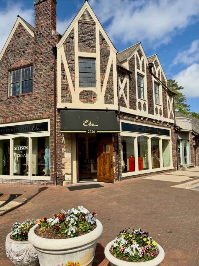 Your Guide to Mountain Brook's Villages - 7 A brick and stucco retail building with large windows, black awnings, and a wooden door, set on a paved plaza with flower planters in the foreground—an inviting scene typical of Mountain Brook Villages.