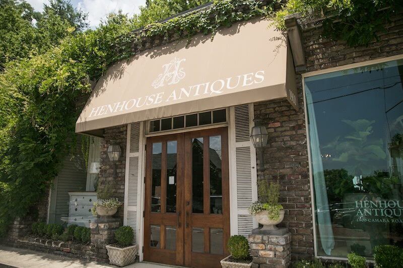 Your Guide to Mountain Brook's Villages - 17 The entrance to Henhouse Antiques in Mountain Brook features a tan awning, double wooden doors, potted plants, and a brick exterior surrounded by greenery—a charming stop in the Villages guide.