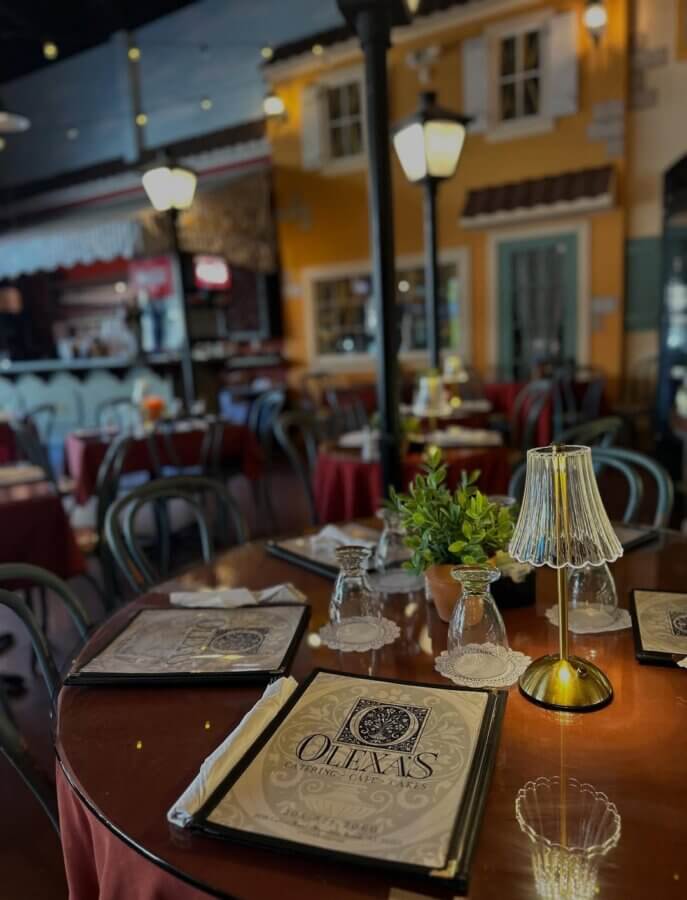 Your Guide to Mountain Brook's Villages - 5 A restaurant interior in Mountain Brook with round tables set with menus, glassware, and small lamps; warm lighting and decor resembling an outdoor European street scene guide guests through charming village vibes.