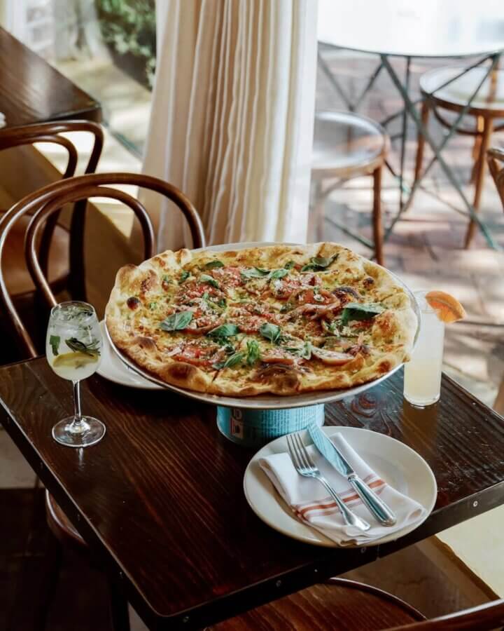 Your Guide to Mountain Brook's Villages - 12 A large pizza topped with vegetables and herbs is served on a raised stand at a Mountain Brook restaurant table, accompanied by two drinks, plates, and cutlery.