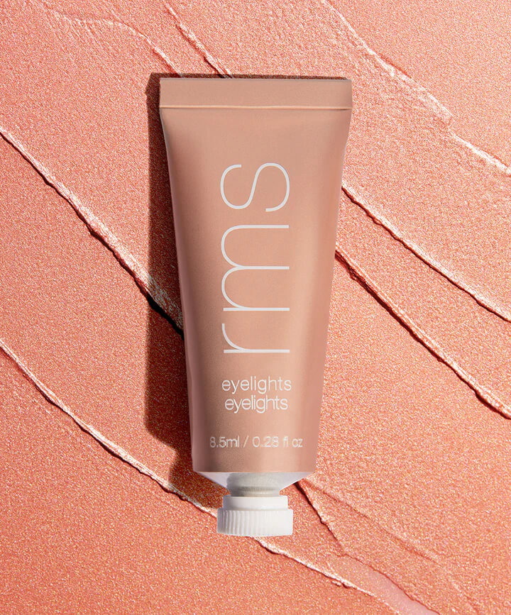 A tube of RMS Eyelights cream eyeshadow lies on a textured, shimmery pink surface.