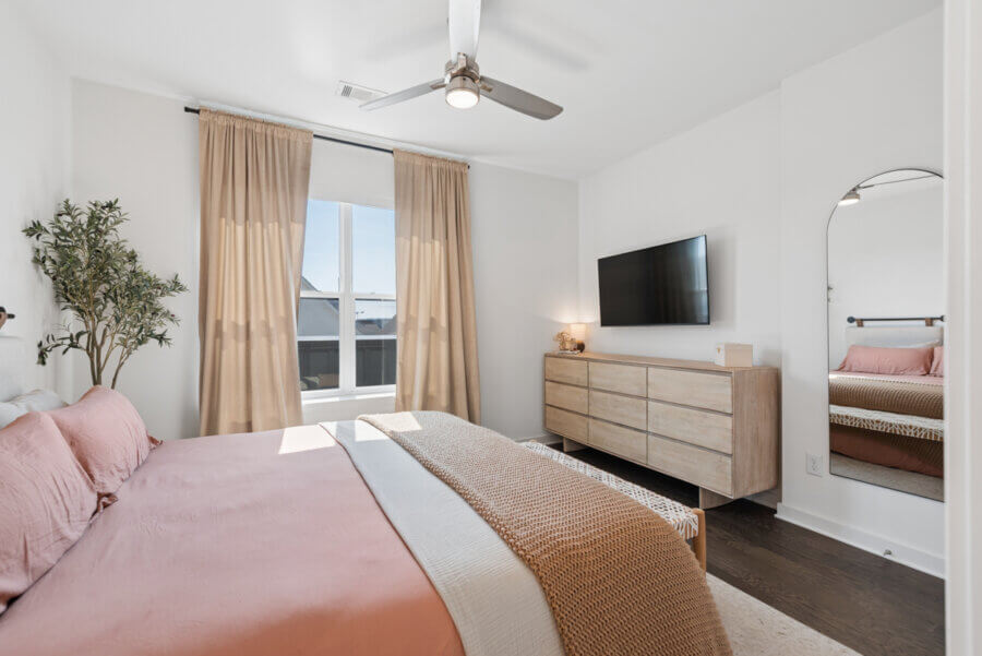 Modern Nashville bedroom with a pink bedding set, wooden dresser, wall-mounted TV, large mirror, potted plant, and a window with beige curtains letting in natural light—perfect for showcasing real estate at 1677 54th Ave N.