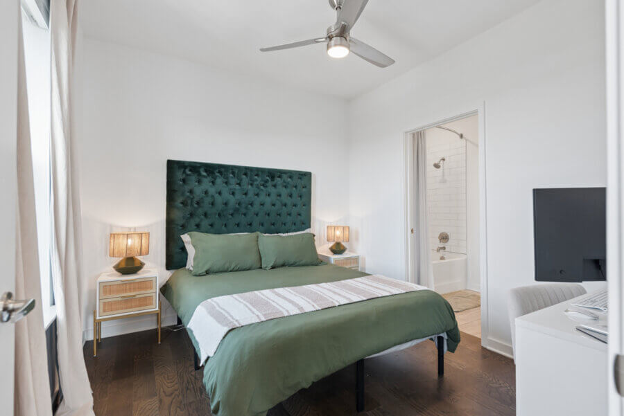 A modern bedroom at 1677 54th Ave N features a green bed, two nightstands with lamps, ceiling fan, and an ensuite bathroom with white tiles and bathtub—perfect for your Nashville real estate dreams.