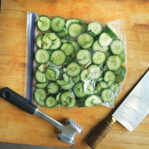 RECIPE: Crushed Garlic and Cucumber Salad - 2 A bag of sliced cucumbers is on a wooden cutting board next to a meat tenderizer and a cleaver.
