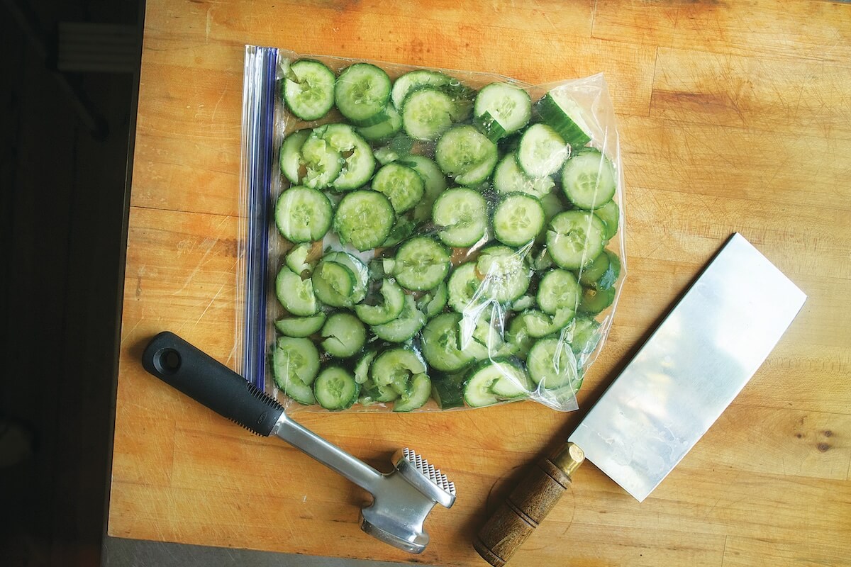 A bag of sliced cucumbers is on a wooden cutting board next to a meat tenderizer and a cleaver.