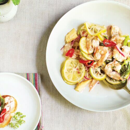 RECIPE: Pickled Shrimp with Coconut Milk - 2 A white plate with shrimp, lemon slices, red chili, and vegetables on a light tablecloth, with a serving spoon and a smaller plate of food beside it.