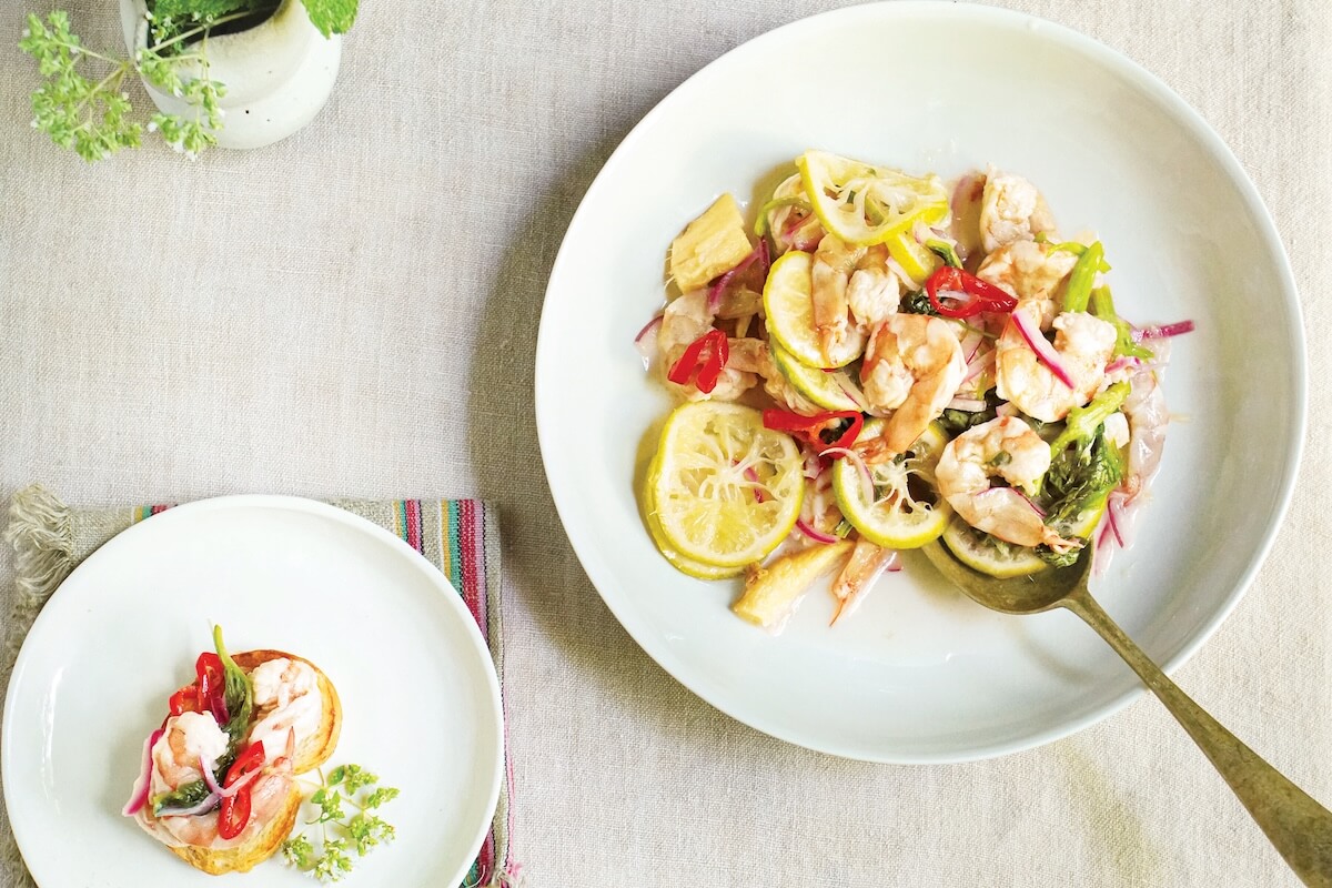 A white plate with shrimp, lemon slices, red chili, and vegetables on a light tablecloth, with a serving spoon and a smaller plate of food beside it.