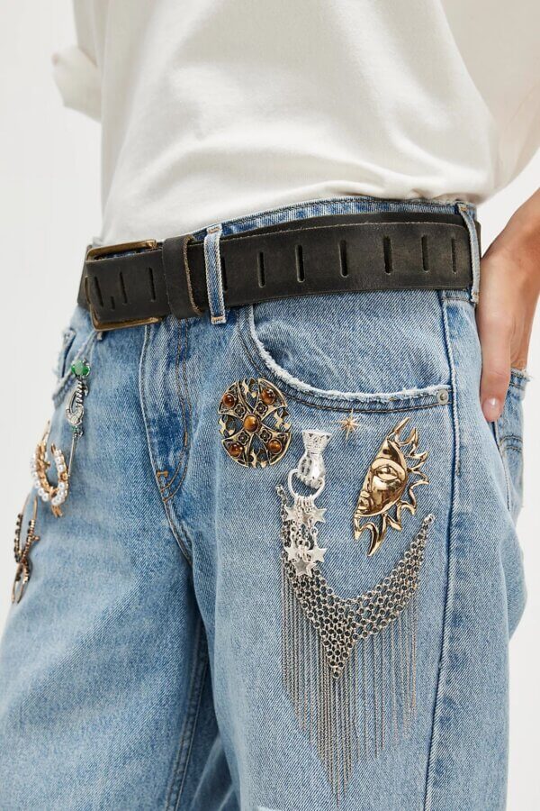 It's the Year of the Brooch! Here's How to Style Them - 8 Close-up of a person wearing light blue denim jeans adorned with metal brooches, chains, and embellishments, styled with a black belt and a crisp white shirt.